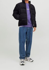 Czarna kurtka puchowa ze sztruksu z wysokim kołnierzem, zapinana na zamek, na fioletowej bluzie, w połączeniu z niebieskimi dżinsami i sportowymi trampkami.