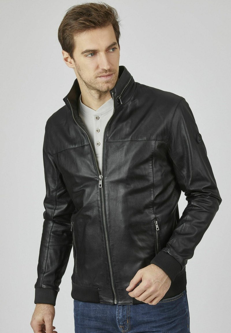 Chaqueta de cuero negra con cremallera frontal, dos bolsillos laterales, puños acanalados y cuello alzado. Textura suave y corte elegante.