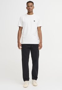 Hvit bomulls-T-skjorte med en liten svart logo på brystet, kombinert med løse svarte bukser og lyse sneakers.