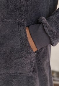Tesut de fâșie gri cu o textură moale, având un buzunar frontal și manșete dantelate. Mânecele au un accent gri mai închis la încheietura mâinii.