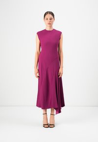 Donna in piedi che indossa un vestito midi senza maniche magenta con orlo asimmetrico e sandali neri con tacco alto e fasce su uno sfondo bianco.