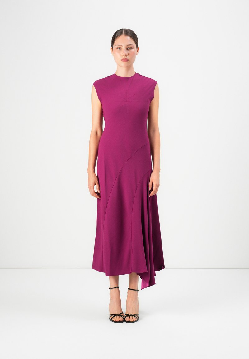 Donna in piedi che indossa un vestito midi senza maniche magenta con orlo asimmetrico e sandali neri con tacco alto e fasce su uno sfondo bianco.