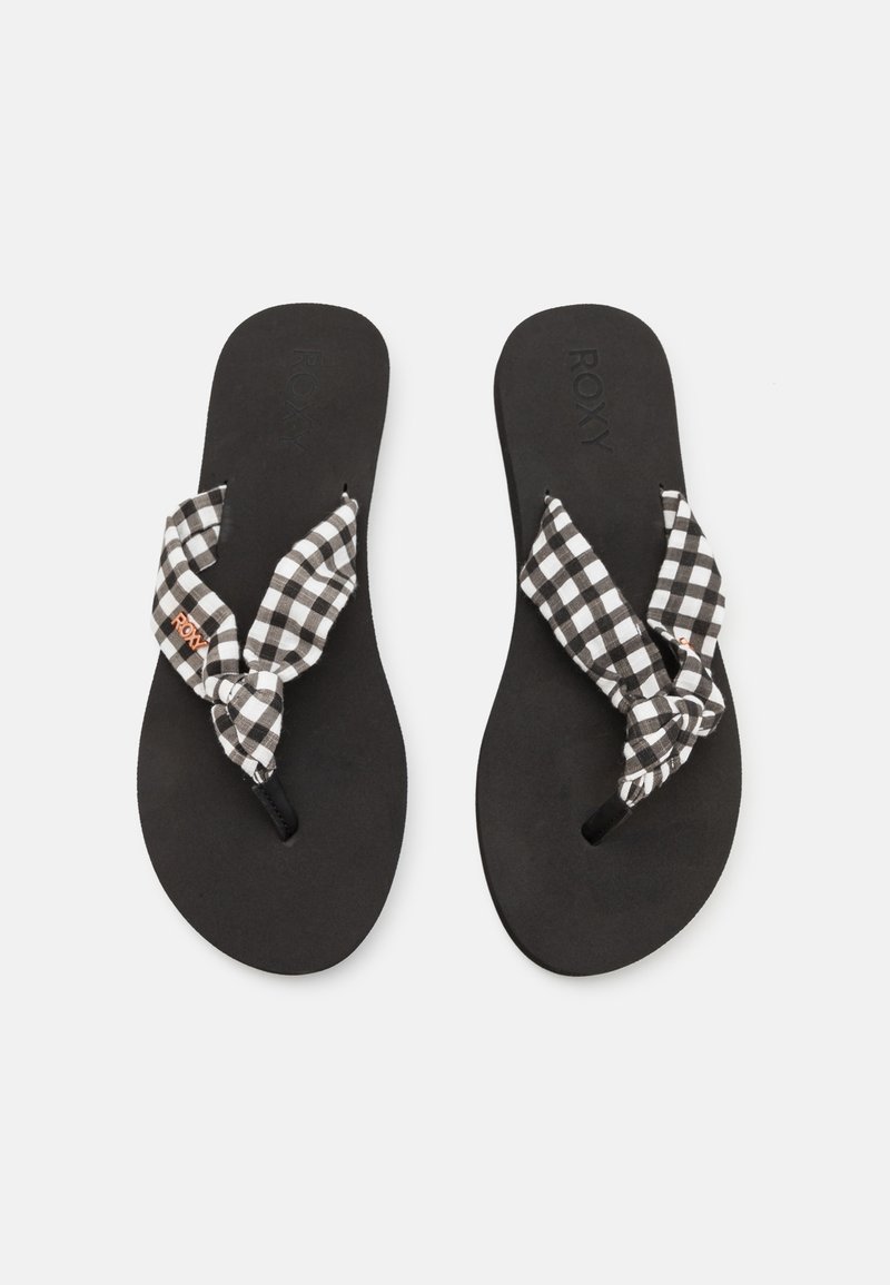 Tongs noires avec des brides en tissu à carreaux noir et blanc doux, nouées en nœuds sur un fond clair uni.