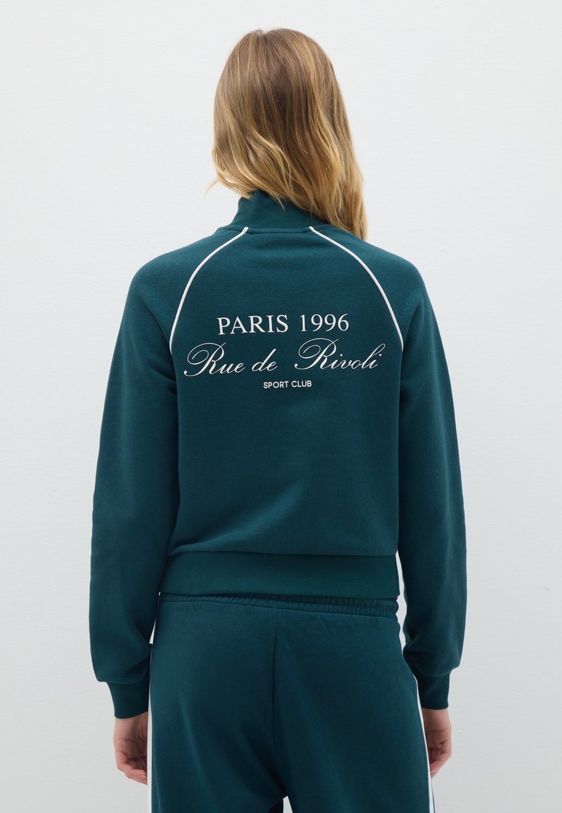 Sudadera de color azul verdoso con acentos blancos; texto "PARÍS 1996 Rue de Rivoli SPORT CLUB" en la parte posterior; puños y dobladillo acanalados; textura de tela suave.