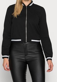 Černá zkrácená bunda se zapínáním na přední zip, žebrovaným límcem a proužkovanými manžetami; doplněná elastickými černými kalhotami z umělé kůže.
