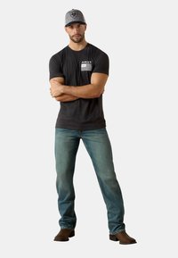 Camiseta negra de manga corta para hombre con la marca "ARIAT" y rayas horizontales, combinada con jeans azul claro y botas marrones. Gorra gris con logo.