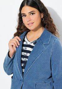 Denim modrý blazer s vykrojeným límcem, zapínáním na jeden knoflík a texturovanou tkaninou. Obsahuje přední kapsy a je vrstvený přes pruhované triko.