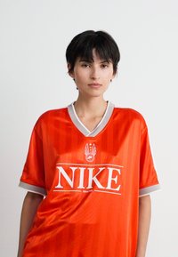 Tânăr cu păr scurt și închis la culoare, îmbrăcat într-un tricou sport Nike supradimensionat de culoare roșie, cu ornamente albe și gri, stând în fața unui fundal alb simplu.