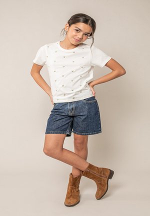 Jong meisje staat met handen in de zij, draagt een wit T-shirt met hartjespatroon, denim shorts en bruine suède enkellaarsjes tegen een beige achtergrond.