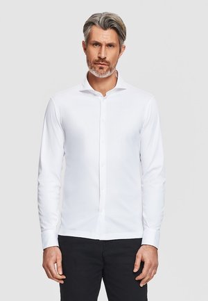 Mężczyzna o siwych włosach i brodzie, ubrany w dopasowaną białą koszulę z guzikami i czarne spodnie, stojący na tle gładkiego jasnego tła.