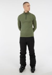 Uomo in piedi con le mani dietro la schiena, indossa una giacca verde oliva con zip, pantaloni neri e scarponi da sci neri su sfondo bianco.