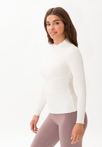 Top bianco a maniche lunghe con texture a coste e scollatura a onda, abbinato a leggings rosa chiaro. Modella in piedi di lato su uno sfondo neutro.
