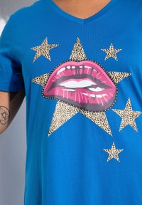 Camiseta azul de cuello en V con un gran gráfico de labios rosas y estrellas de estampado de leopardo. La tela suave y los colores audaces realzan el diseño.
