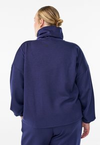 Donna bionda con i capelli raccolti in un bun, che indossa un maglione blu scuro a collo alto e pantaloni abbinati, di spalle contro uno sfondo bianco semplice.