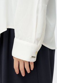 Mano relajada junto a una falda negra, vistiendo una blusa blanca de manga larga con puño abotonado y una pequeña etiqueta rectangular plateada en la manga.