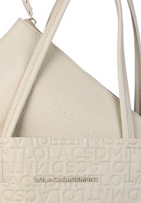 Beige läderbärkasse med en strukturerad yta, som har en framträdande präglad logotyp och släta detaljer. Handtagen är robusta och justerbara.