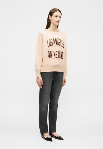 Kvinne står iført en beige sweatshirt med teksten "Los Angeles Anine Bing", mørkegrå jeans og svarte spisse flate sko foran en hvit bakgrunn.