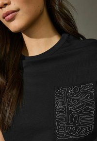 Maglietta nera con tasca caratterizzata da un intricato design di cucitura bianca a linee. Il tessuto appare morbido con un classico scollo rotondo.