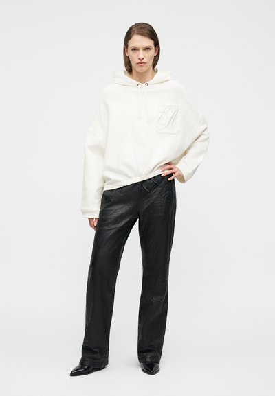 Γυναίκα που φοράει μια φαρδιά λευκή μπλούζα με κουκούλα και μαύρο υφασμάτινο παντελόνι με φαρδιά πόδια, στέκεται με το ένα χέρι στη μέση της.