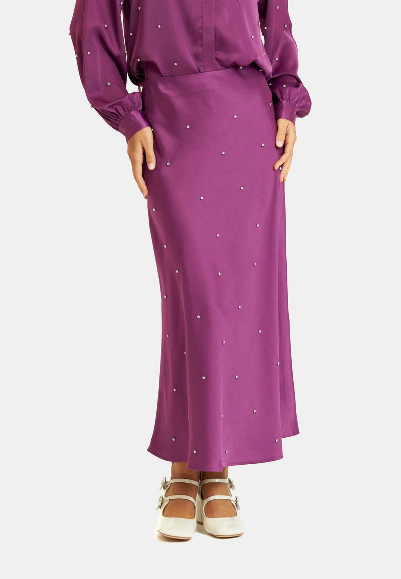 Falda maxi de satén púrpura con tachuelas metálicas dispersas, una silueta elegante y una textura suave, combinada con zapatos blancos de tiras.