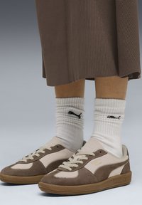 Zapatillas marrones y beige con materiales de ante y cuero, lados texturizados y cordones claros. Combinadas con calcetines blancos de canalé que presentan un logo.
