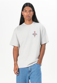 Grå bomull T-skjorte med kort erme, med en fargerik brodert design på venstre bryst. Brukt sammen med blå denimshorts.