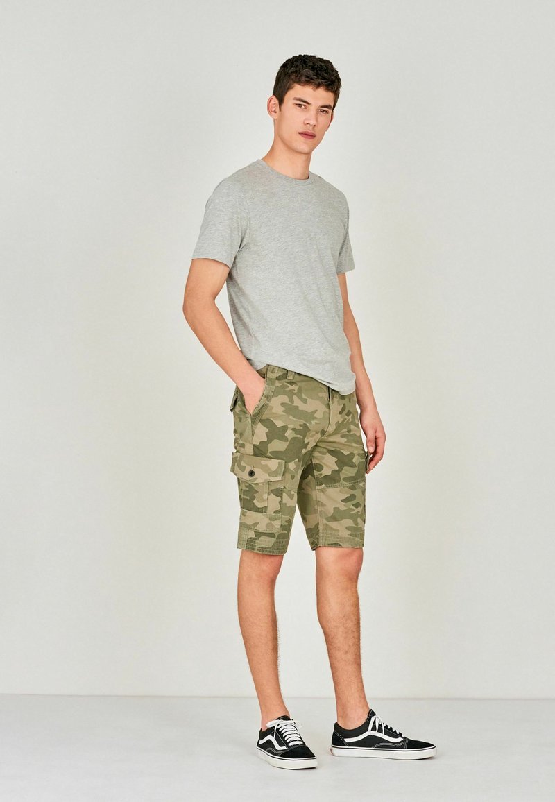 Camiseta gris de manga corta combinada con pantalones cortos de cargo de patrón camuflaje que tienen bolsillos laterales. Zapatillas negras con acentos blancos completan el atuendo.
