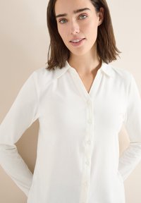 Langärmlige, weiße Bluse mit Knopfleiste, Kragen und glatter Textur. Der Stoff wirkt leicht und weich.