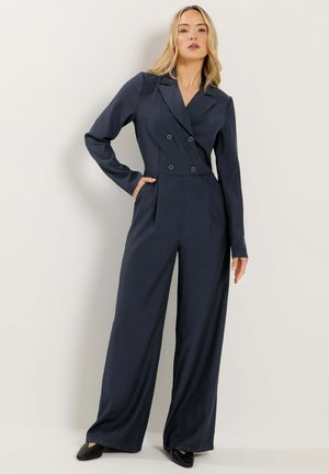Lyshåret kvinde iført en marineblå dobbeltradet blazer-jumpsuit med vide ben og sorte sko, poserer med den ene hånd i lommen.