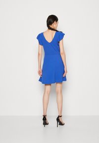 Sininen mekko, jossa on V-mallinen selkä, lyhyet röyhelöhelmaiset hihat, vartalonmyötäinen yläosa, levenevä hame ja sileä pinta. Yhdistetty mustiin korkokenkiin.