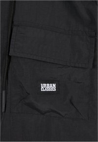 Black fabric jacket featuring a large front pocket with a rectangular Urban Classics logo patch in white. Zip closure along one side.