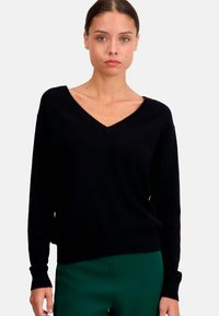Pull noir à col en V avec des manches longues, texture douce, coupe décontractée, poignets et ourlet côtelés, porté avec un pantalon vert.