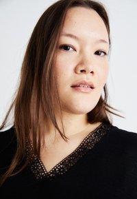 Black top featuring a decorative lace neckline. The fabric is smooth with a relaxed fit. Hair is shoulder-length and styled naturally.