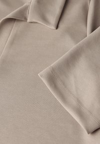 Beige poloshirt met een zachte, gladde textuur, voorzien van een omgeslagen kraag en opgerolde mouwen. Zichtbare stikdetails langs de randen.