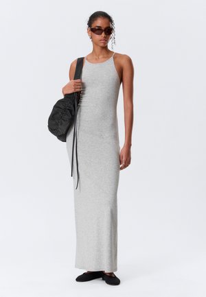 Frau in ärmellosem grauem Maxikleid, schwarzen Ballerinas, Sonnenbrille und mit einer schwarzen, gerafften Umhängetasche vor einem schlichten Hintergrund.