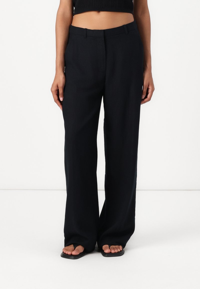 Pantalones amplios negros de tela ligera, con frente plano, bolsillos laterales y una textura suave. Llevados con sandalias de dedo.