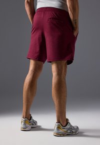 Μπορντό αθλητικές βερμούδες με επίπεδη ζώνη και πίσω τσέπη, φορεμένες με γκρι και κίτρινες αθλητικές κάλτσες μπροστά σε γκρι φόντο.