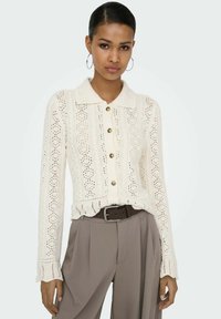 Cardigan en maille crème avec col, présentant une fermeture boutonnée sur le devant, un motif ajouré complexe et un ourlet festonné. Associé à un pantalon taille haute.