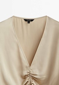 Blouse beige à encolure en V avec un détail froncé au centre et un tissu lisse, montrant la partie supérieure avant et l'étiquette de la marque à l'intérieur du col.