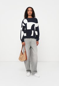 Sudadera navy con patrones abstractos en blanco, combinada con unos pantalones vaqueros de pierna ancha en gris claro y un bolso beige. Zapatillas blancas completan el look.