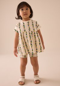 Top chiaro con motivi floreali verticali in verde, viola e giallo; abbinato a pantaloncini a coste con disegni simili; sandali bianchi.