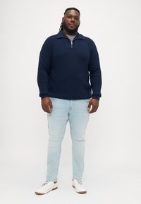 Ναυτικό πλεκτό πουλόβερ με μισό φερμουάρ στον γιακά, συνδυασμένο με ανοιχτό μπλε τζιν και λευκά αθλητικά παπούτσια. Χαλαρή γραμμή με ραβδωτές μανσέτες και πορπη.