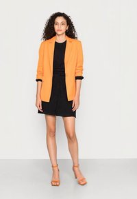 Blazer laranja ajustado sobre um vestido preto, com um tecido texturizado e cintura franzida. Modelo usa sandálias de tiras laranja.