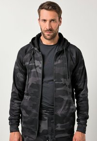 Zwarte camouflage zip-up hoodie met een fleece structuur, voorzien van een trekkoordcapuchon, voorkantzakken en een aansluitend ontwerp. Draagt over een zwart shirt.