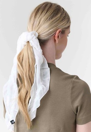 Blonde woman with a brown shirt, hair tied in a low ponytail wrapped with a large white ruffled fabric hair scarf.