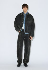 Zwarte spijkerjas met zakken en knoopsluiting, gedragen over een lichtblauw spijkerhemd, gecombineerd met bijpassende zwarte spijkerbroek en zwarte schoenen.