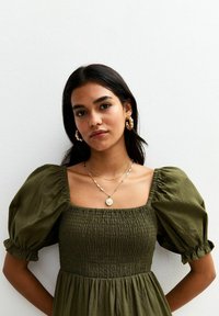 Groene jurk met pofmouwen, vierkante halslijn, gerimpeld lijfje en een vloeiende rok. Gelaagde gouden kettingen en grote hoopoorbellen.