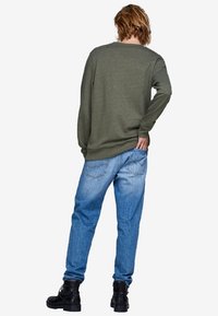 Camisa de manga larga color verde oliva, vista trasera. Combinada con jeans azul claro y botas negras con cordones. Diseño casual con un ajuste relajado.