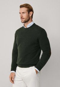 Uomo in maglione verde con toppe sui gomiti sopra una camicia azzurra chiara e pantaloni bianchi, in piedi con una mano in tasca, che guarda di lato.