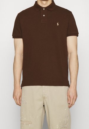 Brun bomulds polo t-shirt med krave, to-knaps lukning og et broderet logo på venstre bryst; parret med lyse slidte bukser.